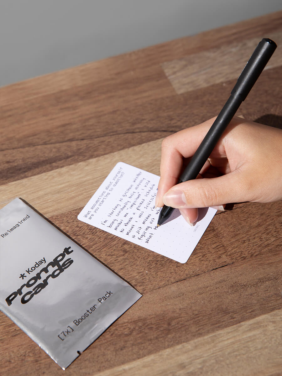 hand writing on a prompt card with a pen, next to a Koday Prompt Cards pack on a wooden surface.